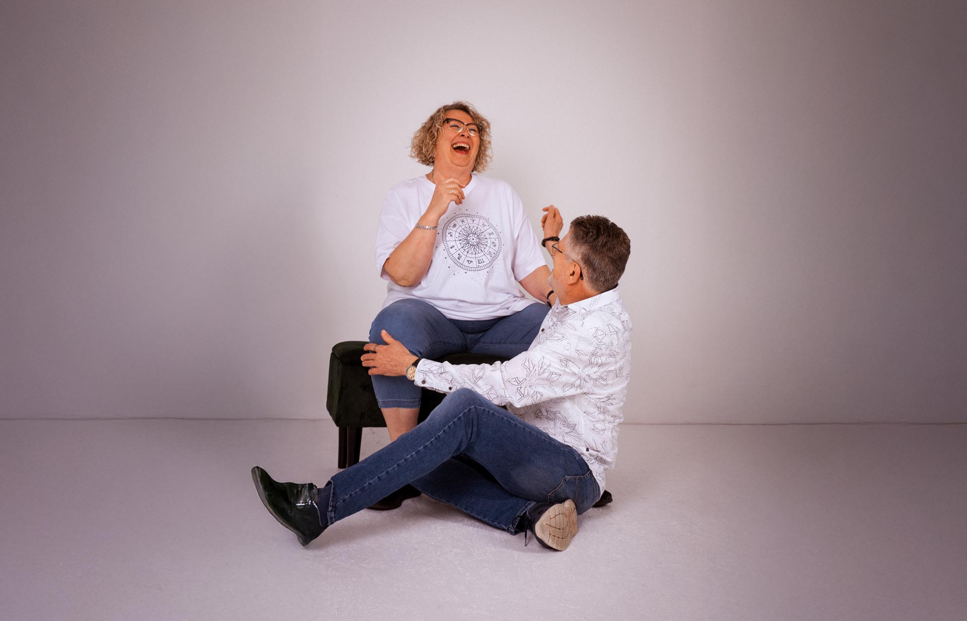 Eine Frau sitzt auf einem Hocker und lacht, während ein Mann vor ihr auf dem Boden sitzt und ebenfalls lacht. Beide scheinen einen spielerischen Moment in einem schlichten, hellen Studio zu genießen.