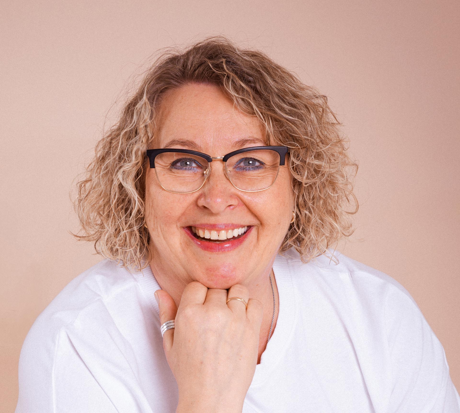 Eine Frau mit lockigem blondem Haar und Brille, die ein weißes t-Shirt trägt, lächelt in die Kamera, während sie vor einem schlichten Hintergrund die Hand unter ihr Kinn legt.