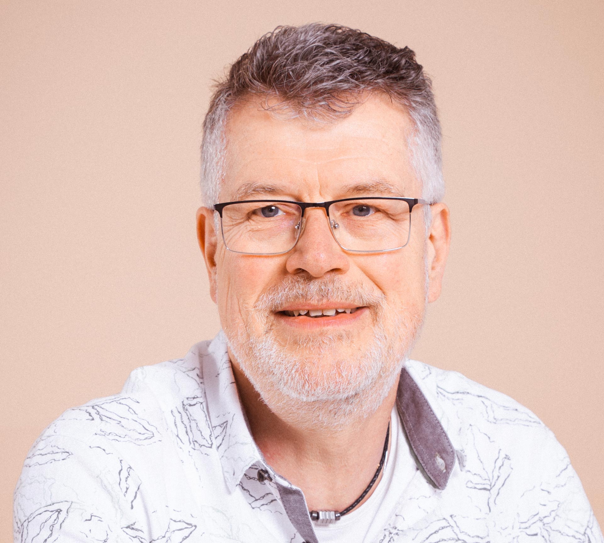 Ein Mann mittleren Alters mit grauem Haar, Brille und kurzem Bart, der ein gemustertes weißes Hemd trägt, lächelt in die Kamera vor einem neutralen Hintergrund.